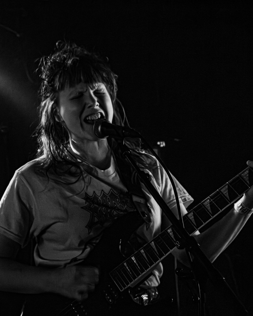 A musician with long hair passionately singing into a microphone while playing an electric guitar, dressed in a graphic t-shirt. The image is in black and white, with dynamic lighting highlighting their intense expression.