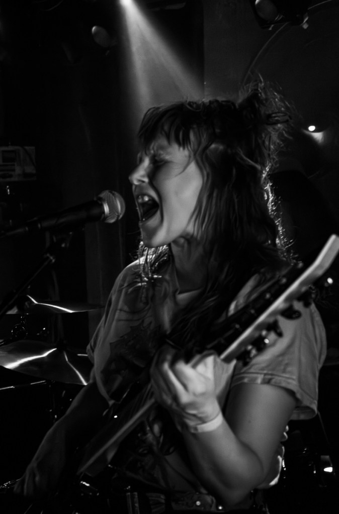 A female singer passionately performing while playing an electric guitar on stage, with dramatic lighting casting shadows and highlights.