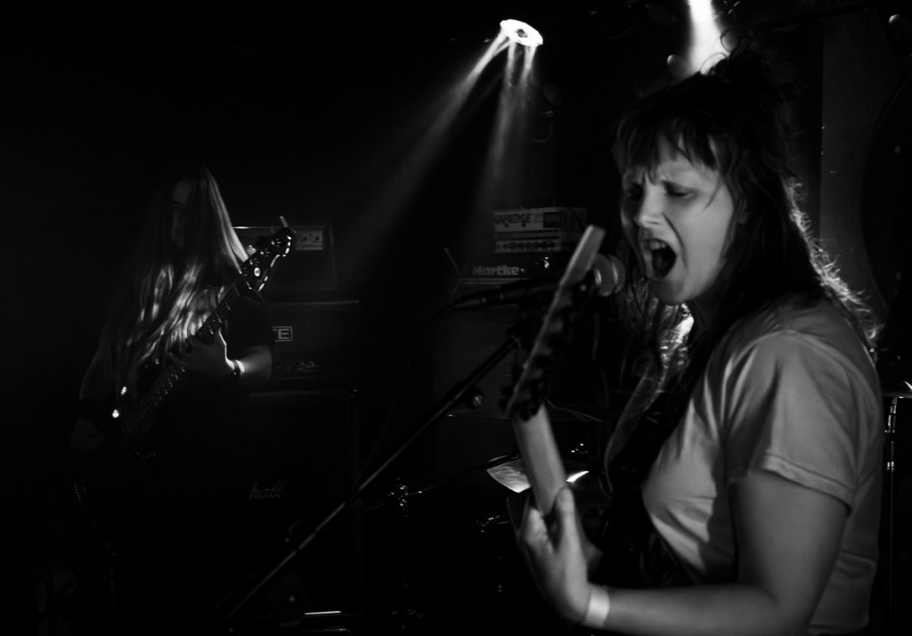 Black and white image of a female vocalist passionately singing into a microphone, while a male guitarist with long hair plays in the background, with stage lights creating a dramatic atmosphere.