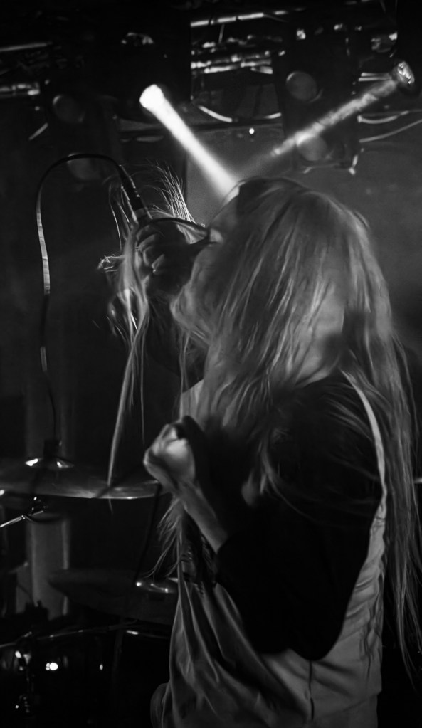 A musician passionately performing on stage with long hair, holding a microphone, surrounded by dramatic lighting effects.