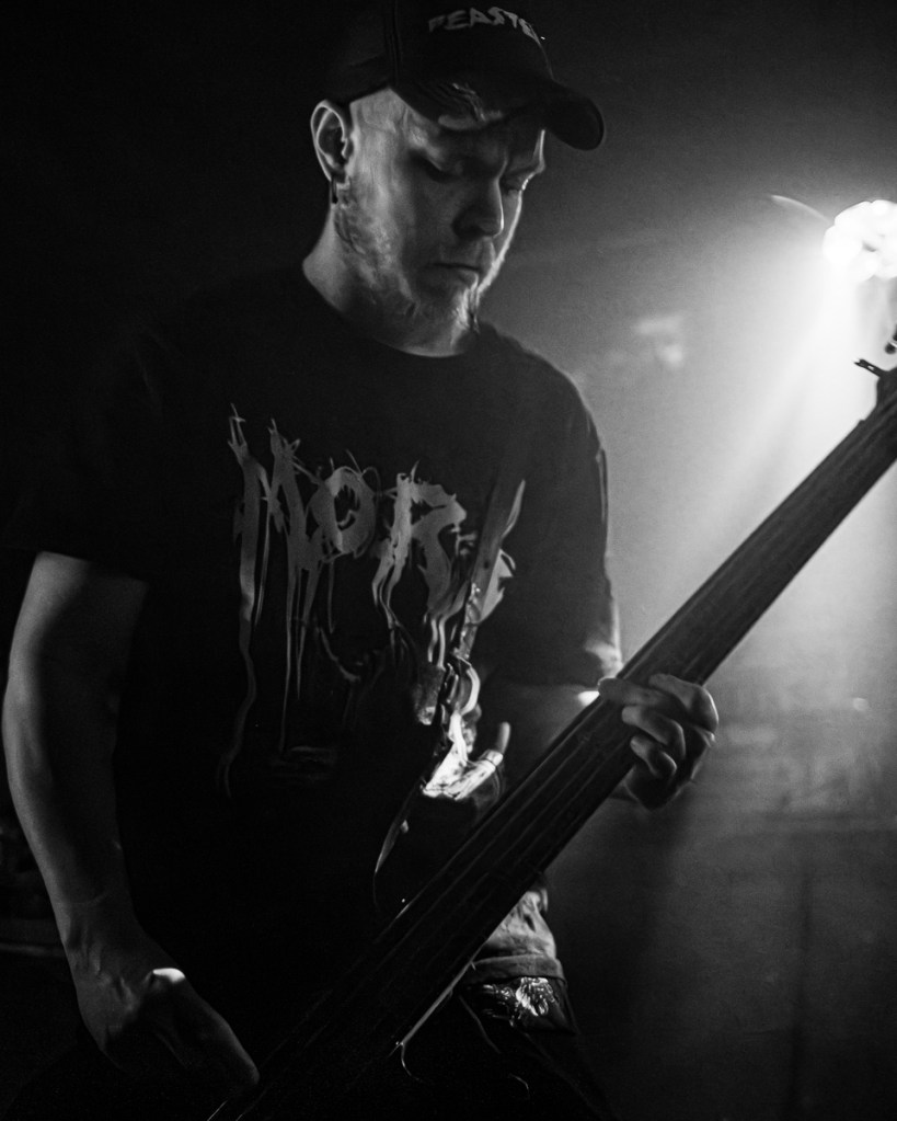 A musician performing on stage, playing a bass guitar, wearing a dark graphic t-shirt and a cap, in a low-light setting.