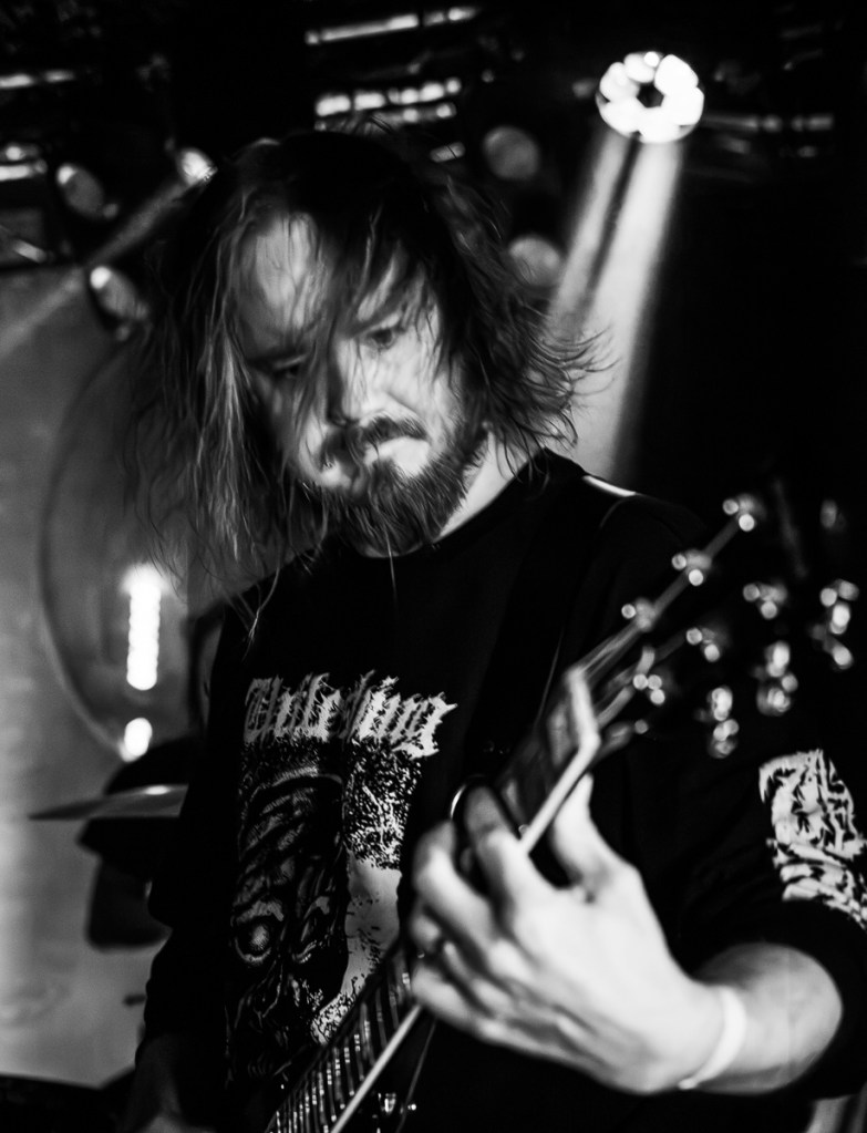 A musician passionately playing an electric guitar on stage, captured in a dynamic moment with long hair and a beard, wearing a black graphic sweatshirt in a concert setting.