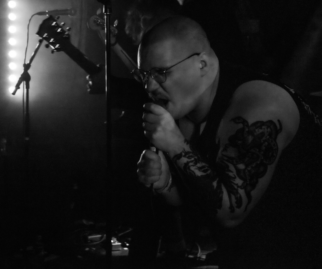 A male vocalist with glasses passionately sings into a microphone on stage, showcasing body tattoos, with blurred band members and instrumental equipment in the background under dim lighting.