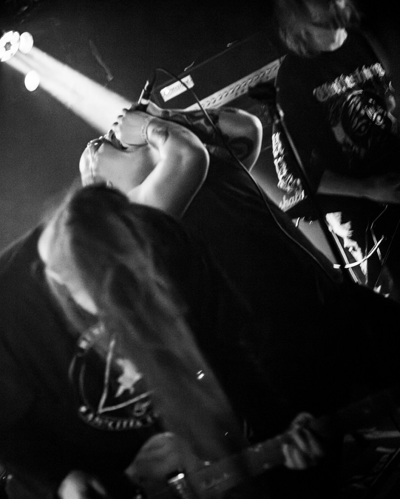 A black-and-white photo of a live music performance featuring a vocalist passionately singing into a microphone while another musician plays a guitar, with dramatic lighting and motion blur.