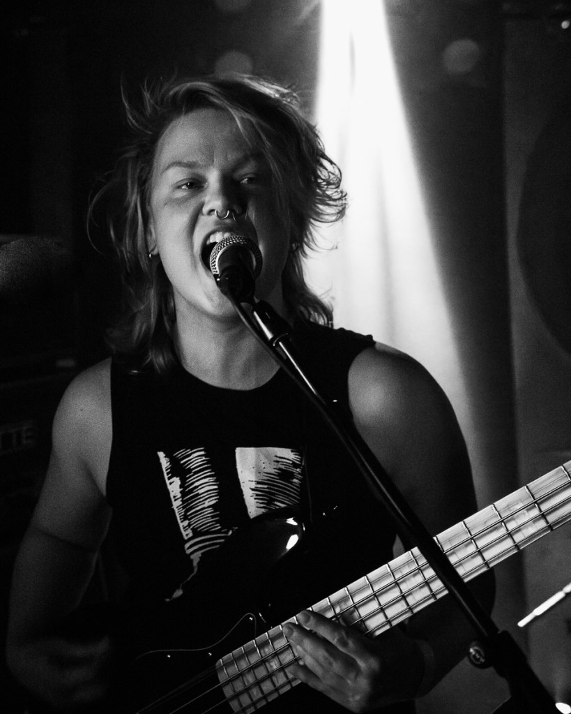 A musician passionately singing into a microphone while playing a bass guitar, with dramatic lighting and a black-and-white color scheme.