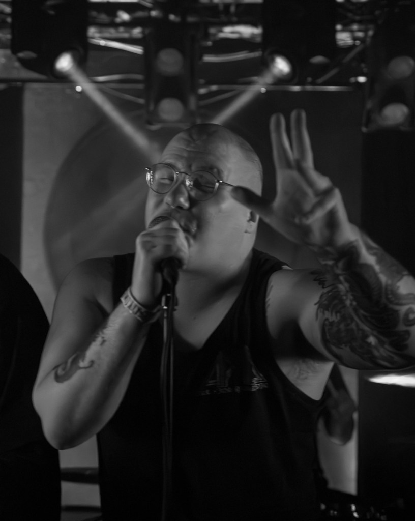 A male singer with glasses passionately performing on stage, holding a microphone and gesturing with his hand. The scene is illuminated by stage lights, creating a dynamic atmosphere.