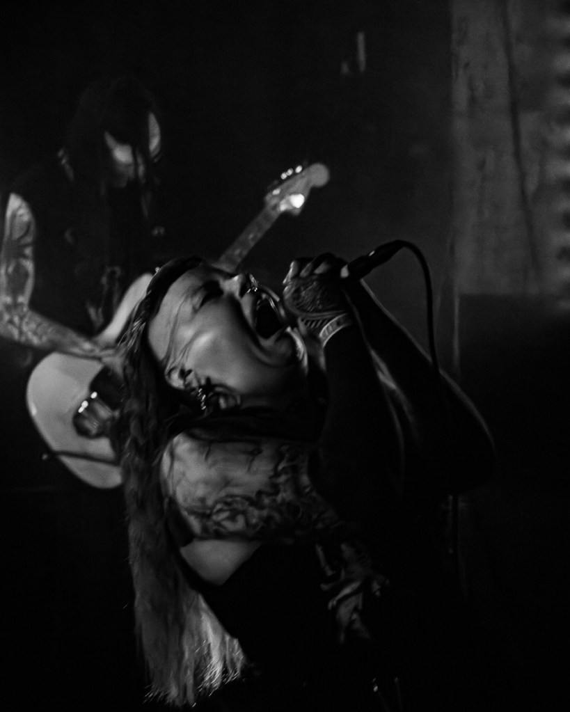 A tattooed singer passionately performing on stage with a microphone, while a guitarist is playing in the background in a dimly lit venue.