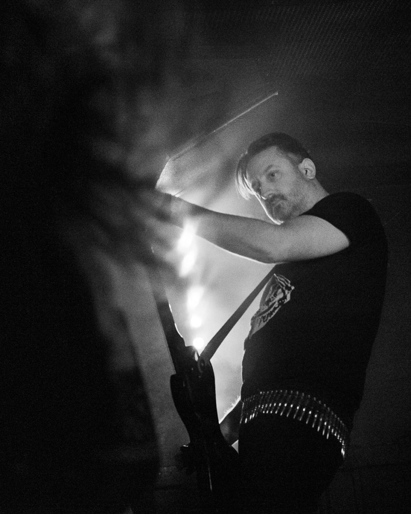 A guitarist performing on stage, captured in black and white with dramatic lighting.