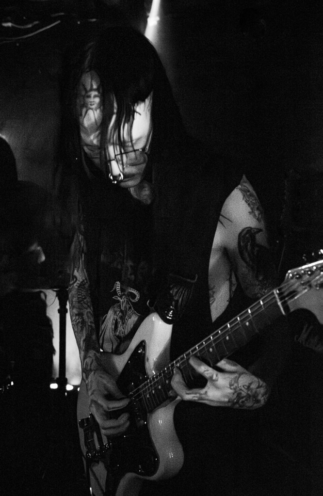 A musician with long dark hair and tattoos performs passionately on stage, playing an electric guitar under dim lighting.
