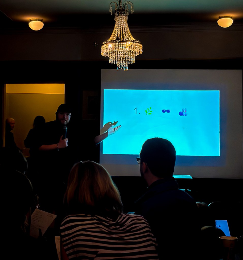 A speaker presenting in front of a projection screen displaying icons, while an audience listens attentively. A chandelier is visible in the background.